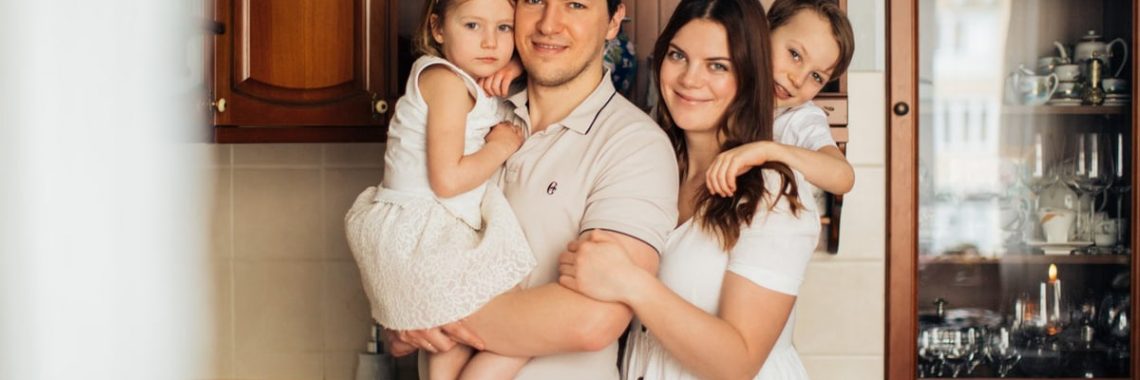 Satisfied man with daughter and woman with son embracing embracing each other while standing against wooden cupboard in kitchen and looking at camera