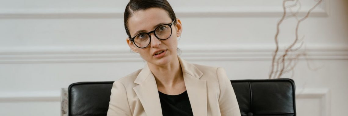 A woman in a beige blazer sitting with eyeglasses and notebook indoors.